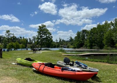 2 kayaks, one red and one lime green, on the grass in front of Merritt's Mill Pond on a sunny blue sky day.